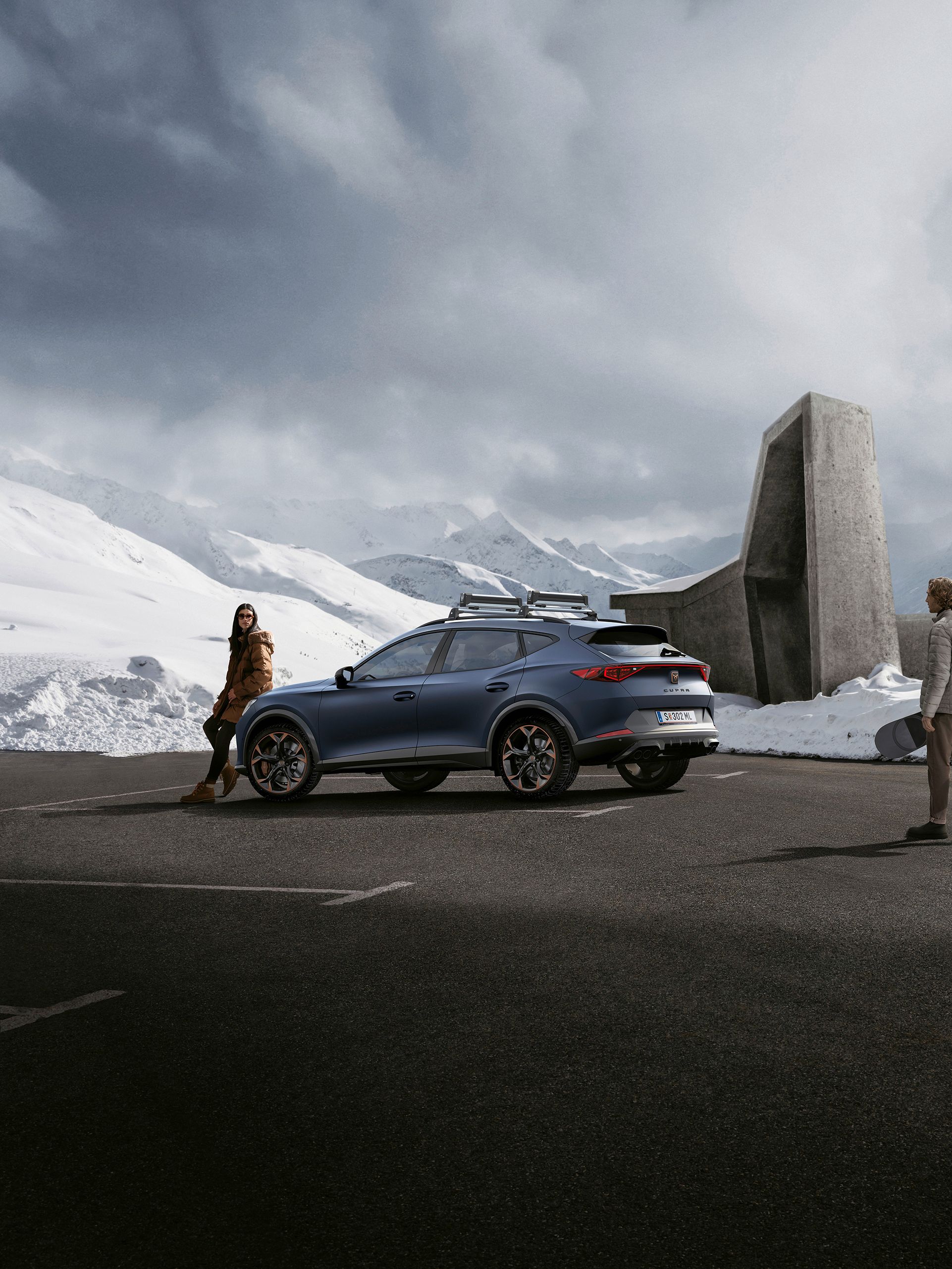 Eine Frau lehnt an einem CUPRA Formentor auf einem winterlichen Parkplatz in den Bergen. Ein Mann mit Snowboard bewegt sich in Richtung des Fahrzeugs.