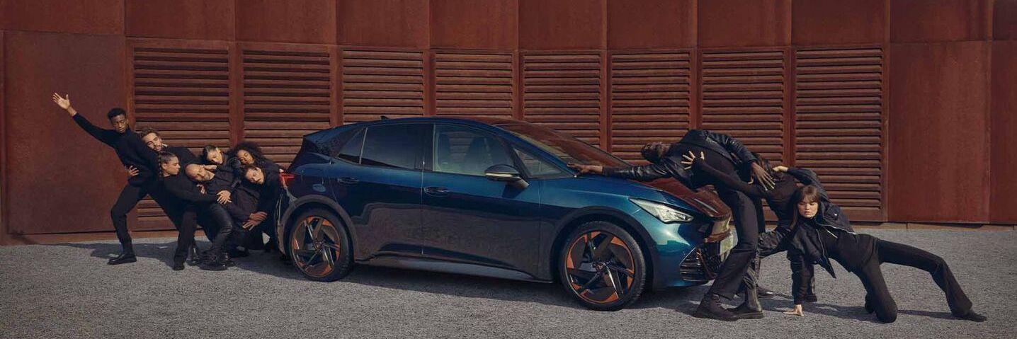 Ein blauer CUPRA Born steht auf einem Platz, Menschen performen neben dem Fahrzeug.