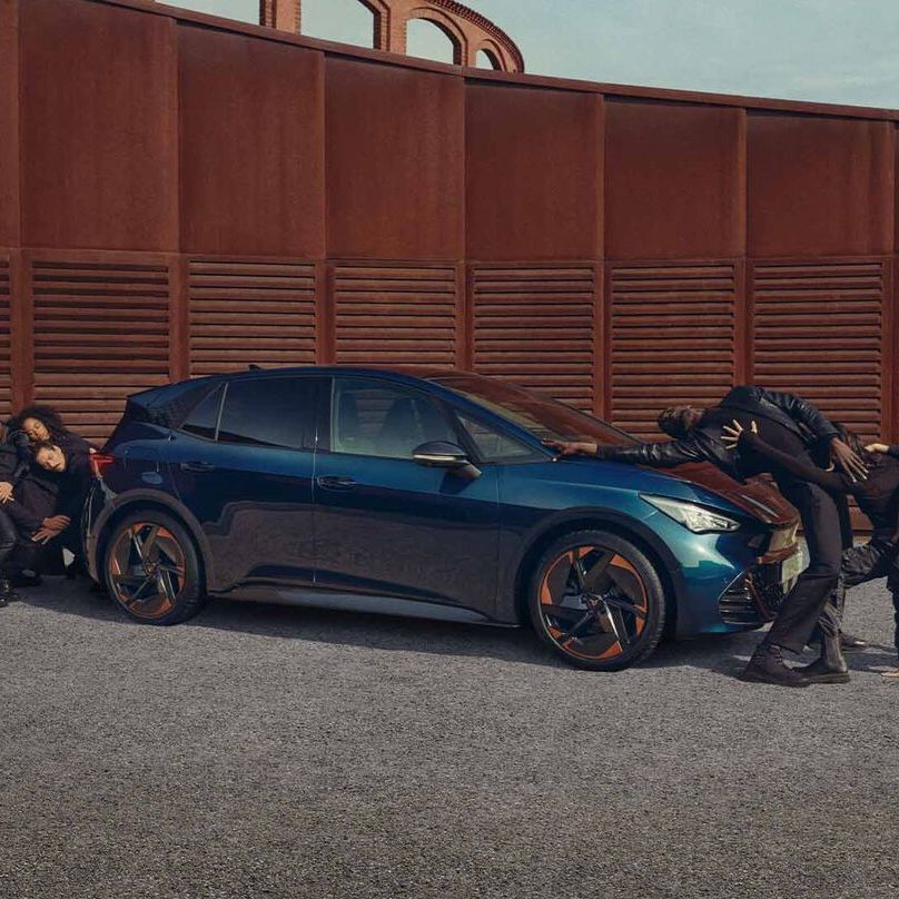 Ein blauer CUPRA Born steht auf einem Platz, Menschen performen neben dem Fahrzeug.