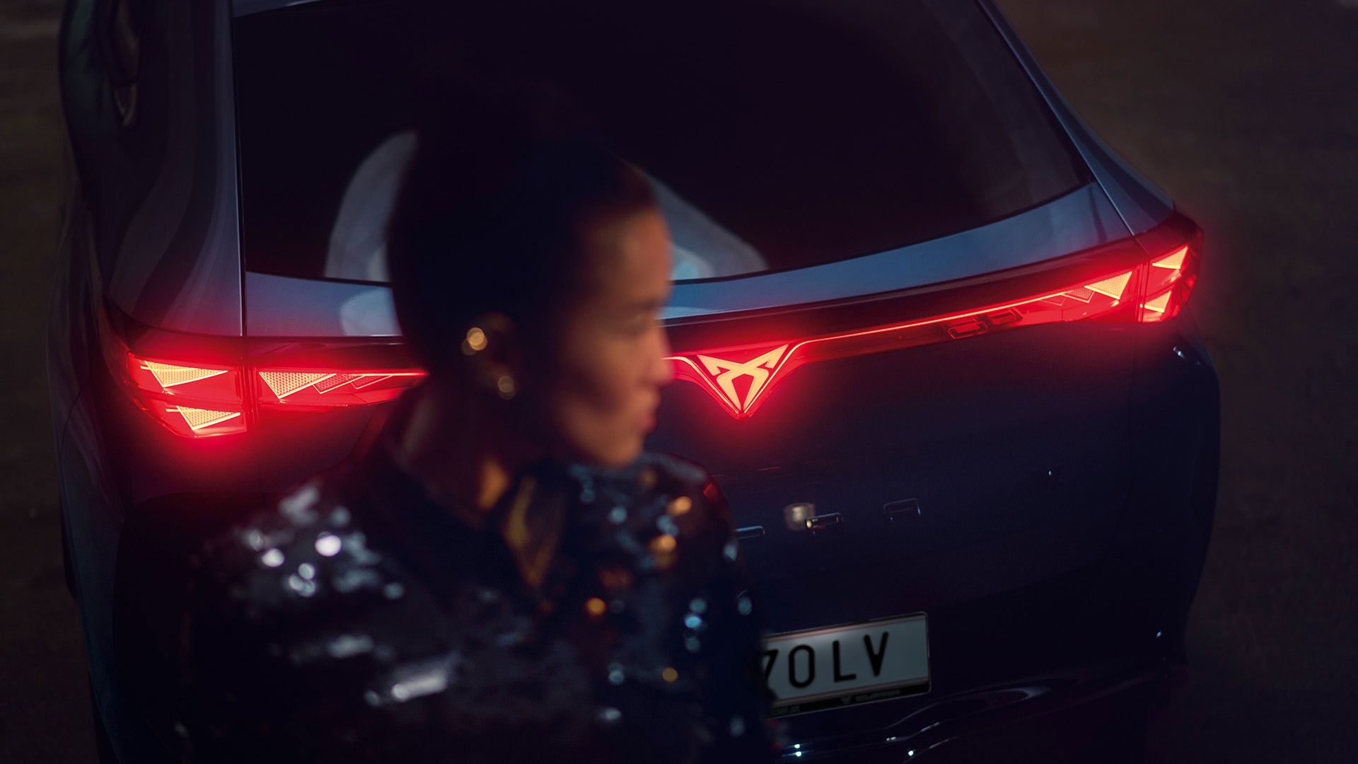 Detailaufnahme vom Heck eines CUPRA Tavascan mit durchgehender Heckbeleuchtung, eine Frau steht neben dem Fahrzeug.