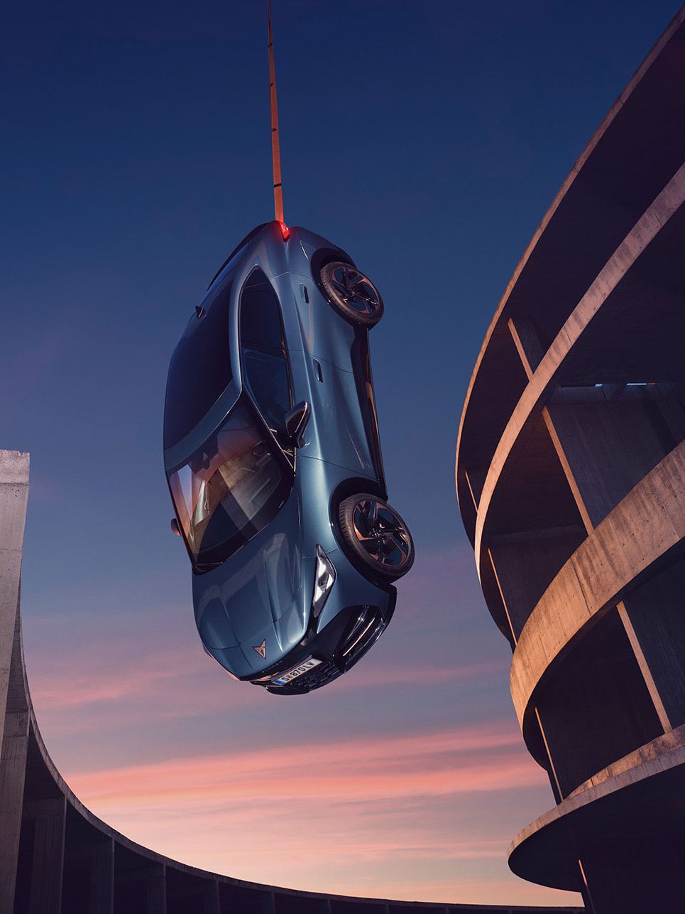 Blauer CUPRA Tavascan erscheint aufgehängt, eine Frau steht daneben auf einem Gebäude.