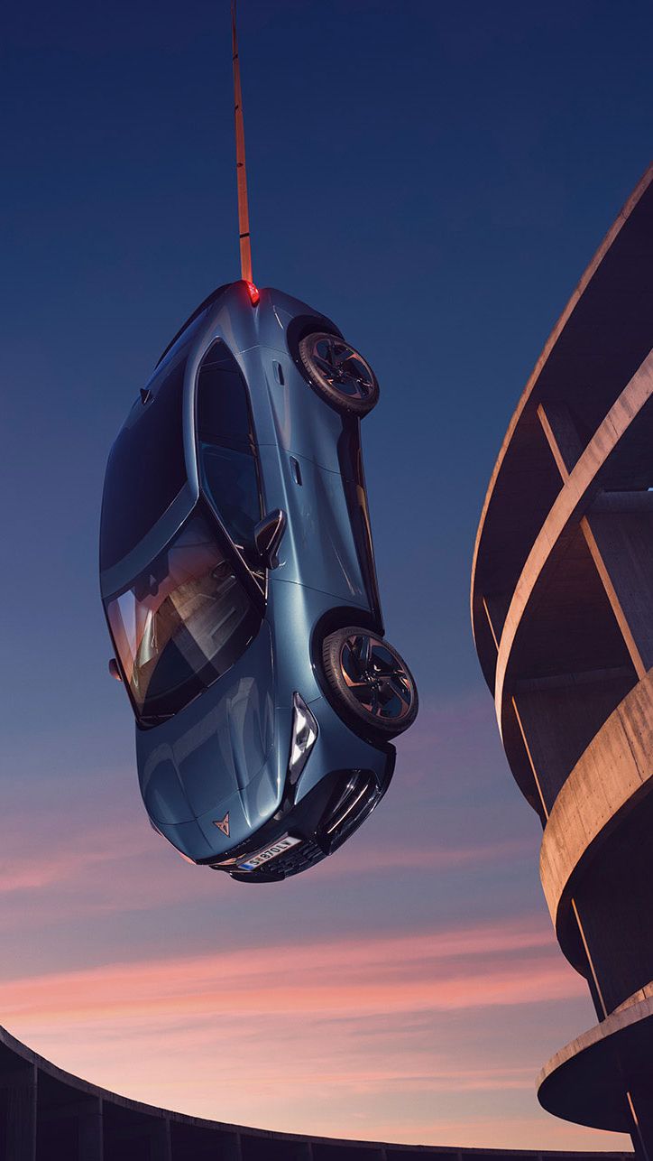 Blauer CUPRA Tavascan erscheint aufgehängt, eine Frau steht daneben auf einem Gebäude.