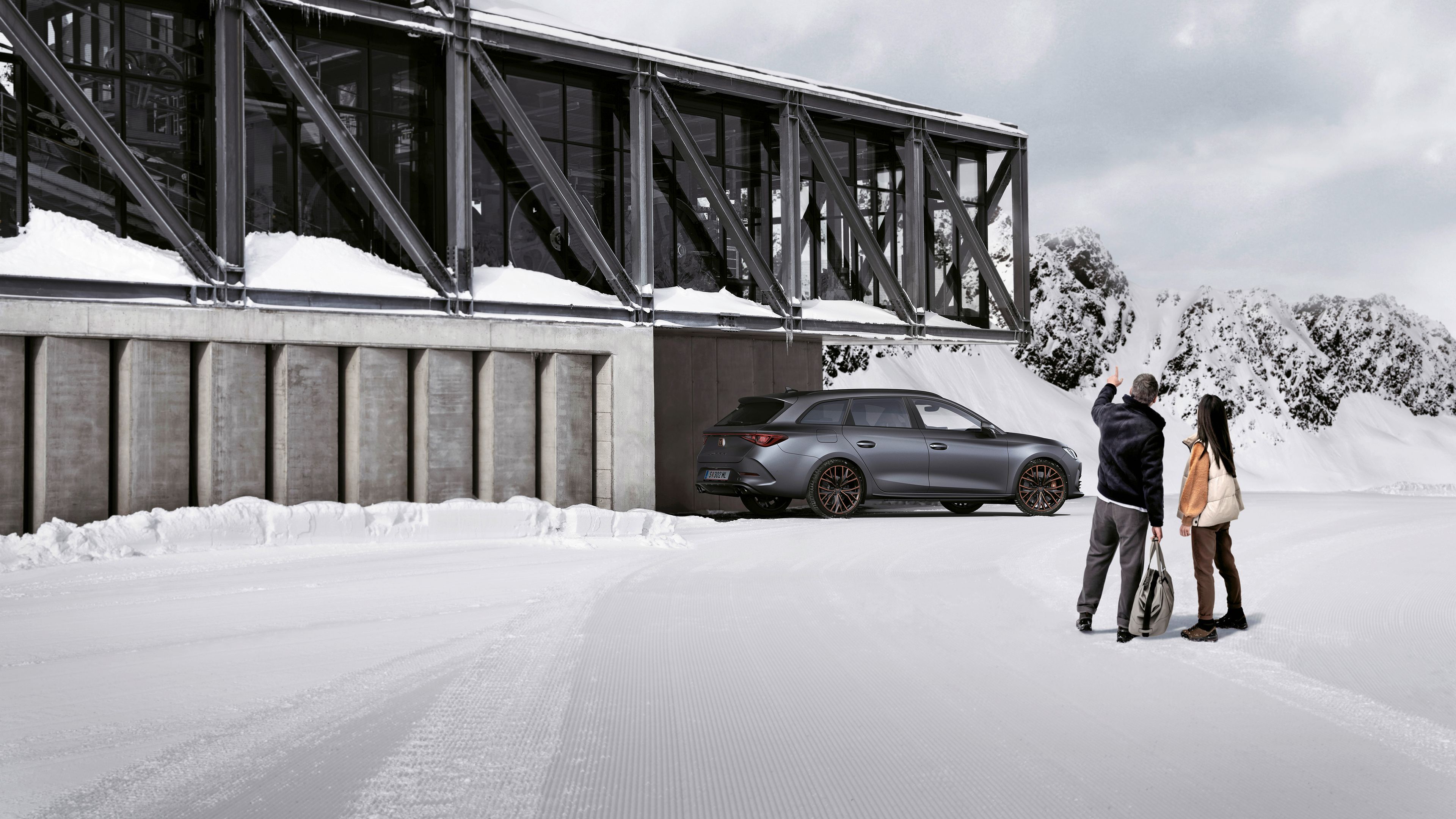 Ein grauer CUPRA Leon SP Kombi parkt vor einer Seilbahnstation in einer winterlichen Berglandschaft. Ein Mann und eine Frau blicken in Richtung des Fahrzeugs.