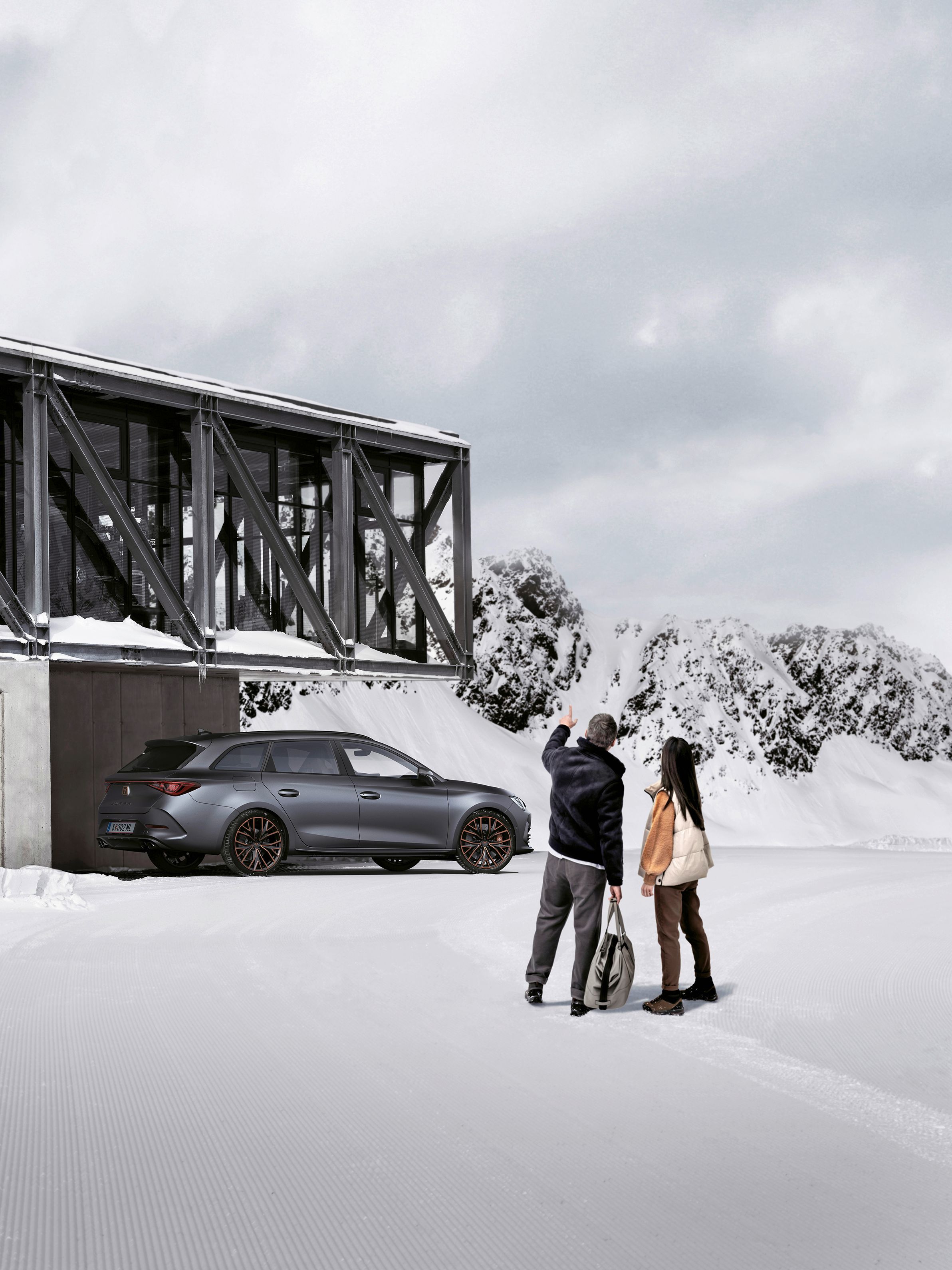 Ein grauer CUPRA Leon SP Kombi parkt vor einer Seilbahnstation in einer winterlichen Berglandschaft. Ein Mann und eine Frau blicken in Richtung des Fahrzeugs.